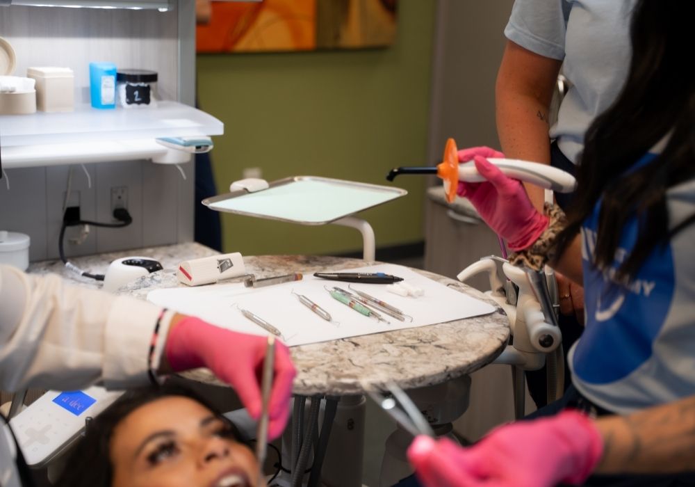 A dentist and assistant, both wearing pink gloves, prepare dental tools and a curing light while a patient lies in the dental chair with their mouth open.