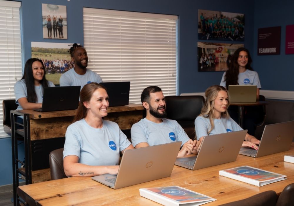 Six people in matching blue shirts sit at desks with laptops in a classroom setting, smiling and looking forward. Several books are on the desks, and colorful posters hang on the walls.