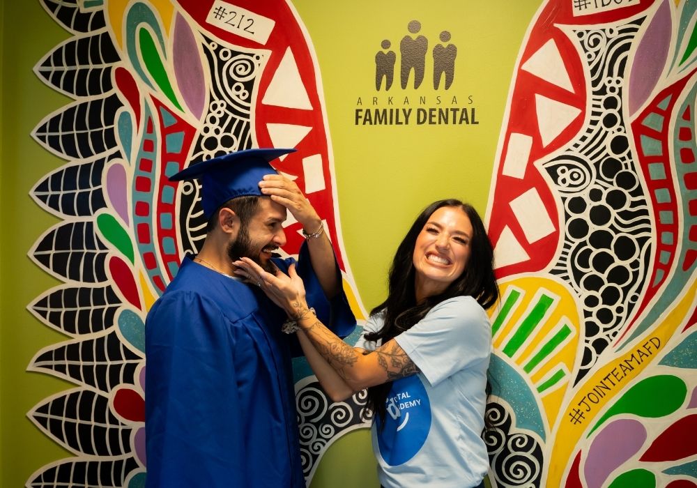A man in a blue graduation gown and cap stands by a smiling woman in a light blue shirt, playfully touching his face. They pose in front of a colorful wing mural at Arkansas Family Dental.