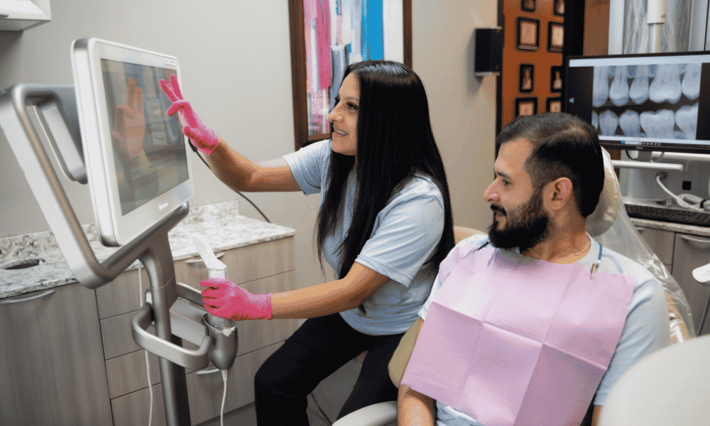 About Avid (6) A dental professional wearing pink gloves shows a male patient dental images on a screen in a modern dental office. The patient is seated, wearing a pink bib, and both are smiling.