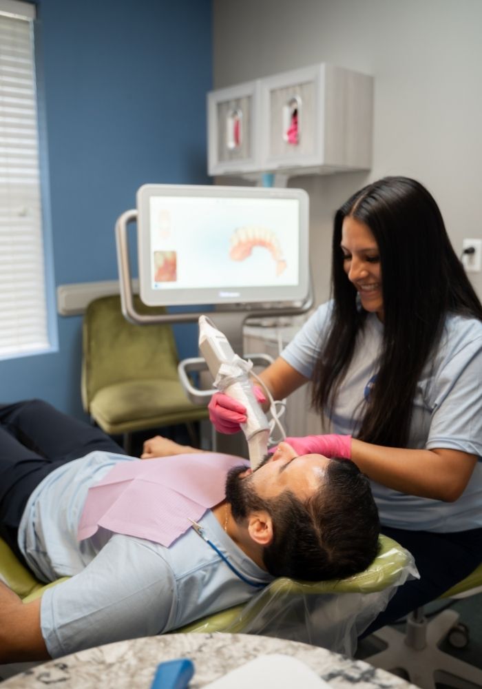 Avid Dental (13) A dental hygienist in gloves uses a digital scanner on a patients teeth while he reclines in a dental chair, with a monitor displaying a 3D dental image in the background.