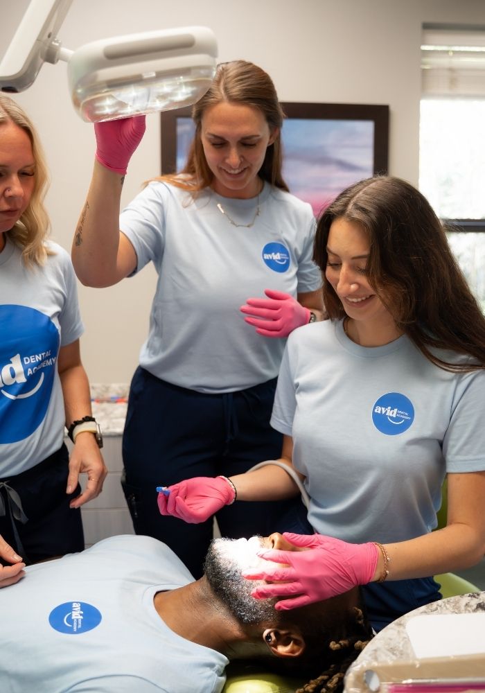Avid Dental (14) Three dental professionals in light blue shirts and pink gloves work on a patient lying in a dental chair under a bright exam light. The atmosphere appears friendly and clinical.