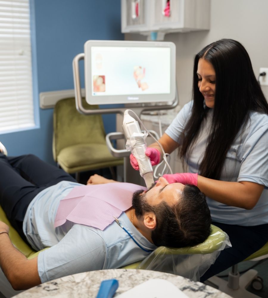 A dental professional uses a handheld scanner to examine a patients teeth in a modern dental office. The patient is reclined in the dental chair, wearing a bib, while the dentist smiles and wears pink gloves.