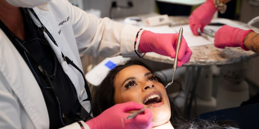 A woman lies in a dental chair with her mouth open while two dental professionals wearing pink gloves examine her teeth using dental instruments.