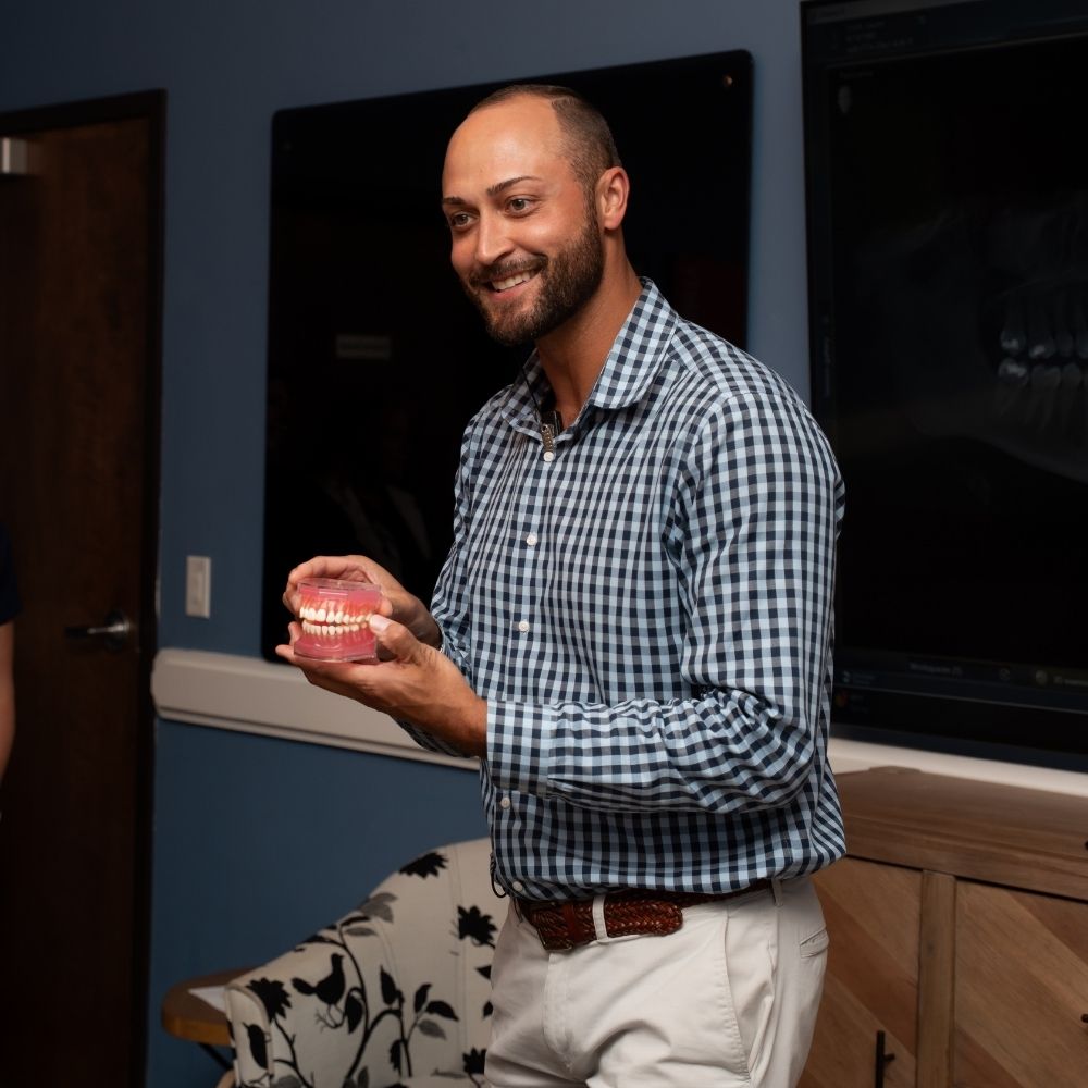 A man in a checkered shirt smiles while holding a dental model of teeth, standing in a room with a chair and dark display boards on the wall behind him.