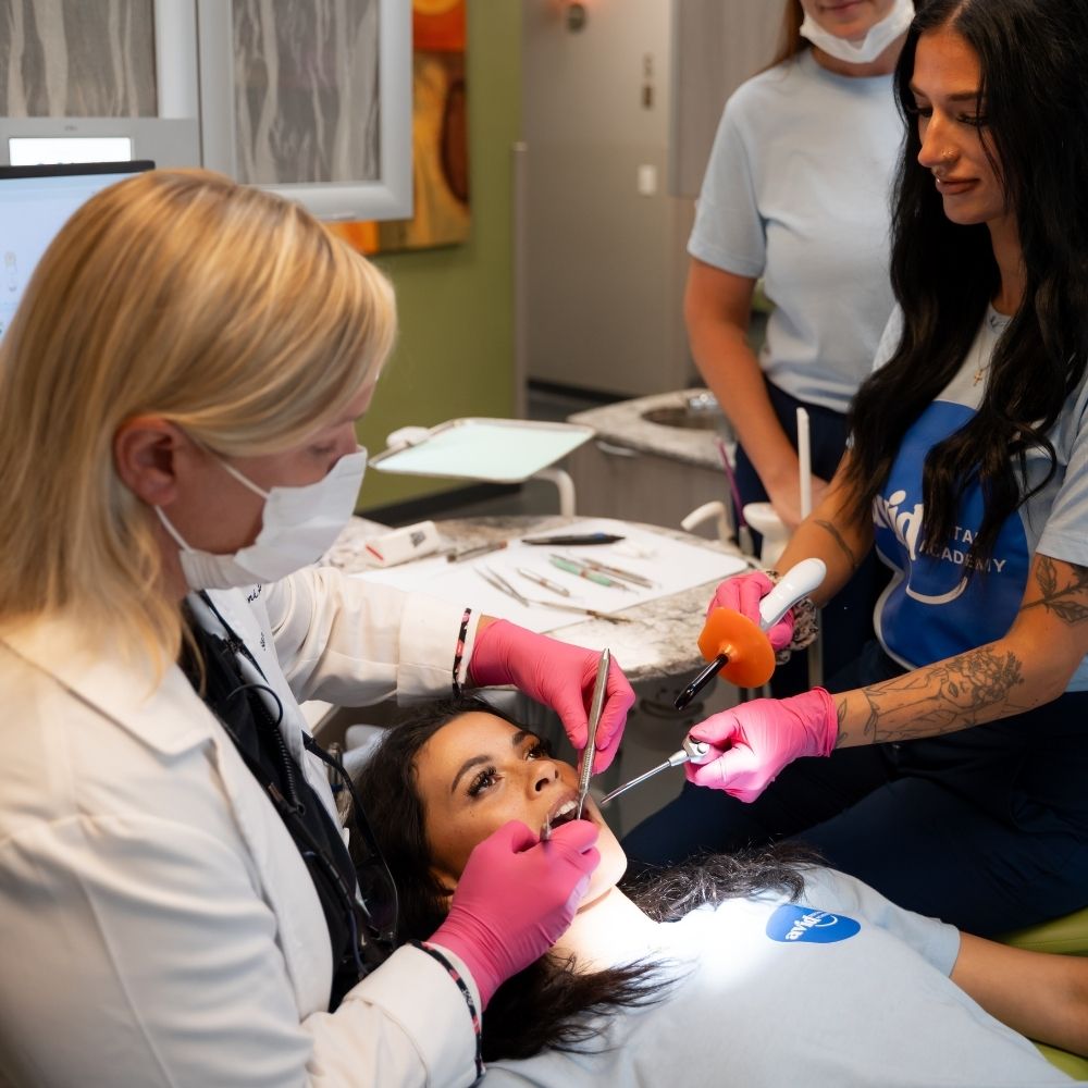 A dental professional examines a womans teeth while two assistants observe. The patient lies in a dental chair. All staff wear gloves and uniforms; one holds a dental tool and another uses a light.