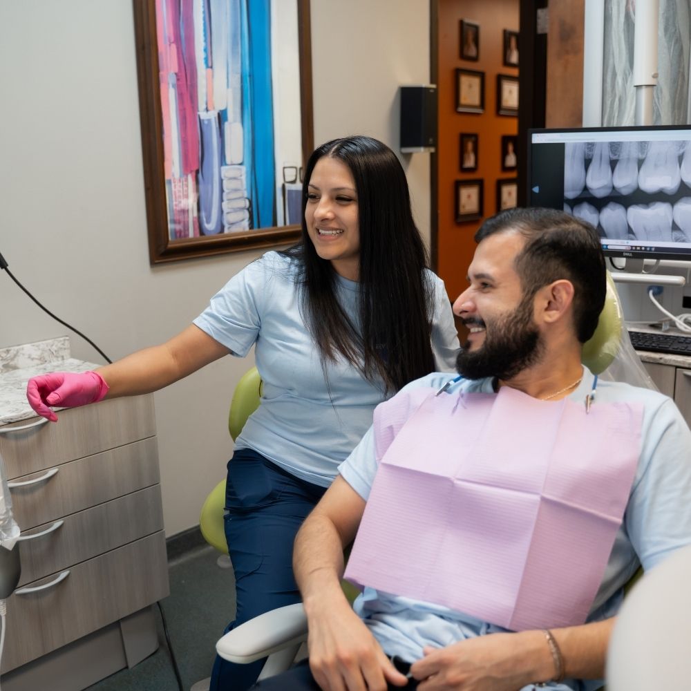 A dentist wearing pink gloves and a male patient smile in a dental office. The patient sits in the chair with a pink bib, while dental X-rays are displayed on a monitor in the background.
