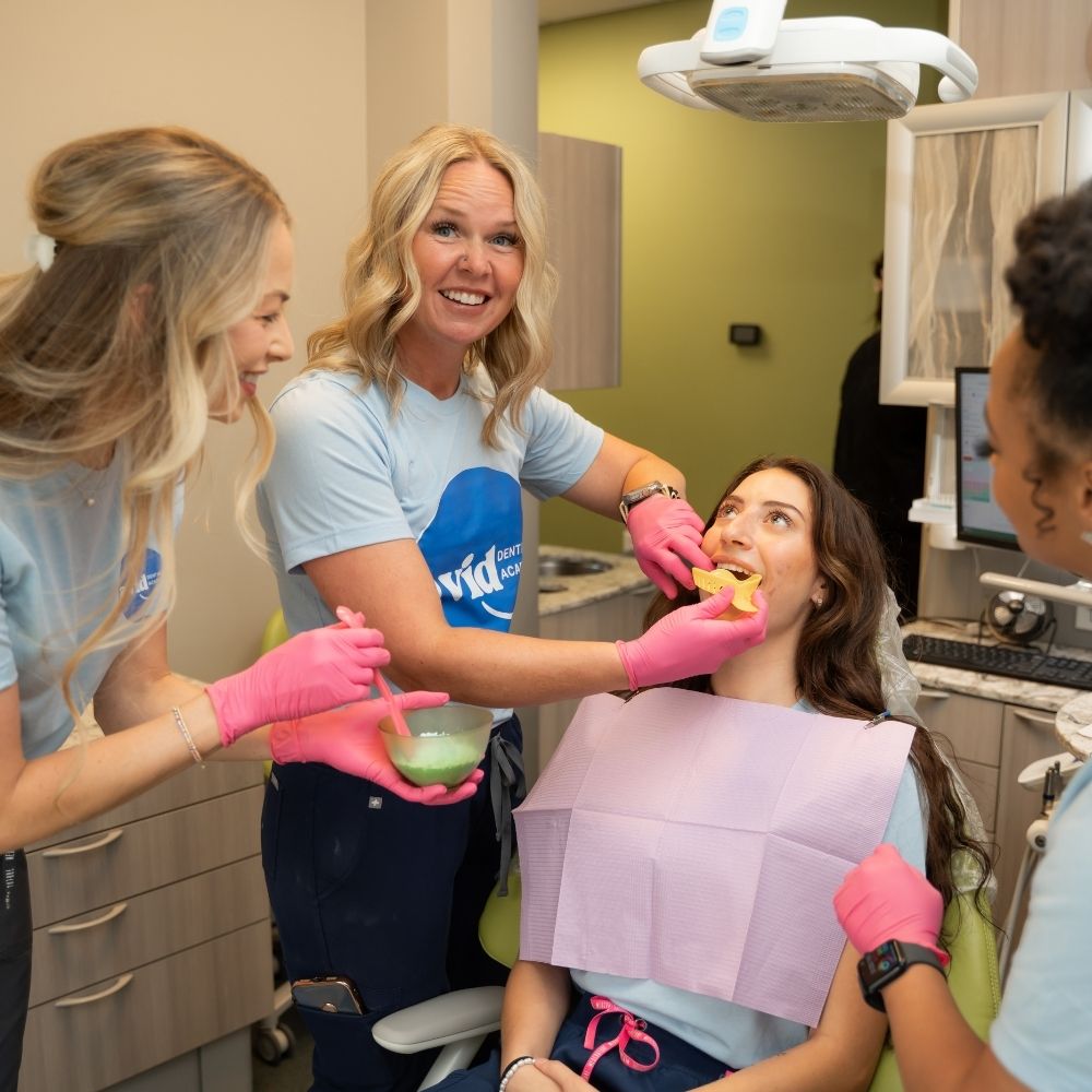 A dental hygienist smiles while applying treatment to a seated patients teeth. Two other staff members watch and smile. All wear gloves, and the patient wears a protective bib in a modern dental office.
