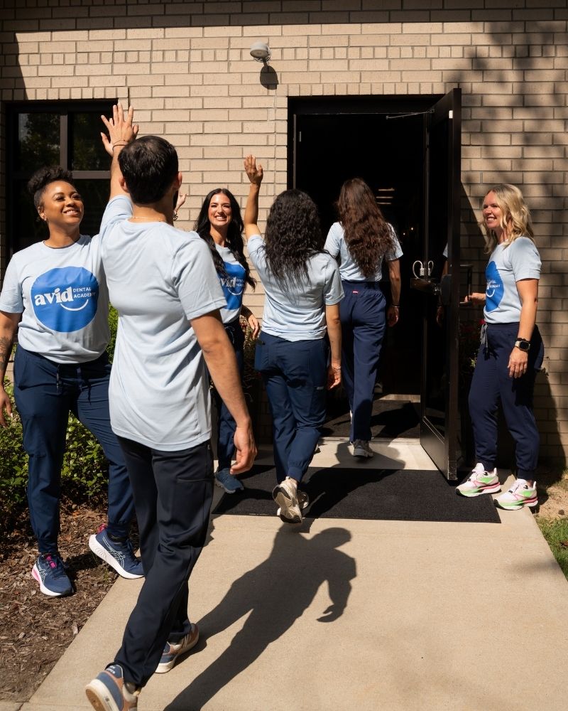 A group of people wearing matching avid shirts high-five and smile as they enter a building together on a sunny day.