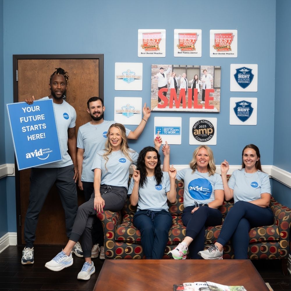 Six people in matching light blue shirts sit and stand in a bright office, smiling and pointing up at awards and a SMILE sign on the wall. One person holds a sign that says, YOUR FUTURE STARTS HERE!.