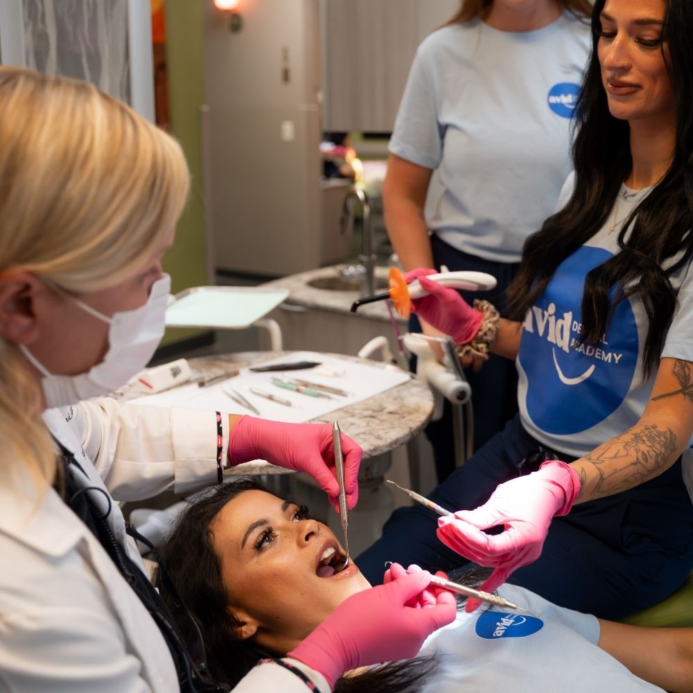 A dental professional in a mask examines a patients mouth while two assistants, all wearing gloves, assist with dental tools in a brightly lit clinic.