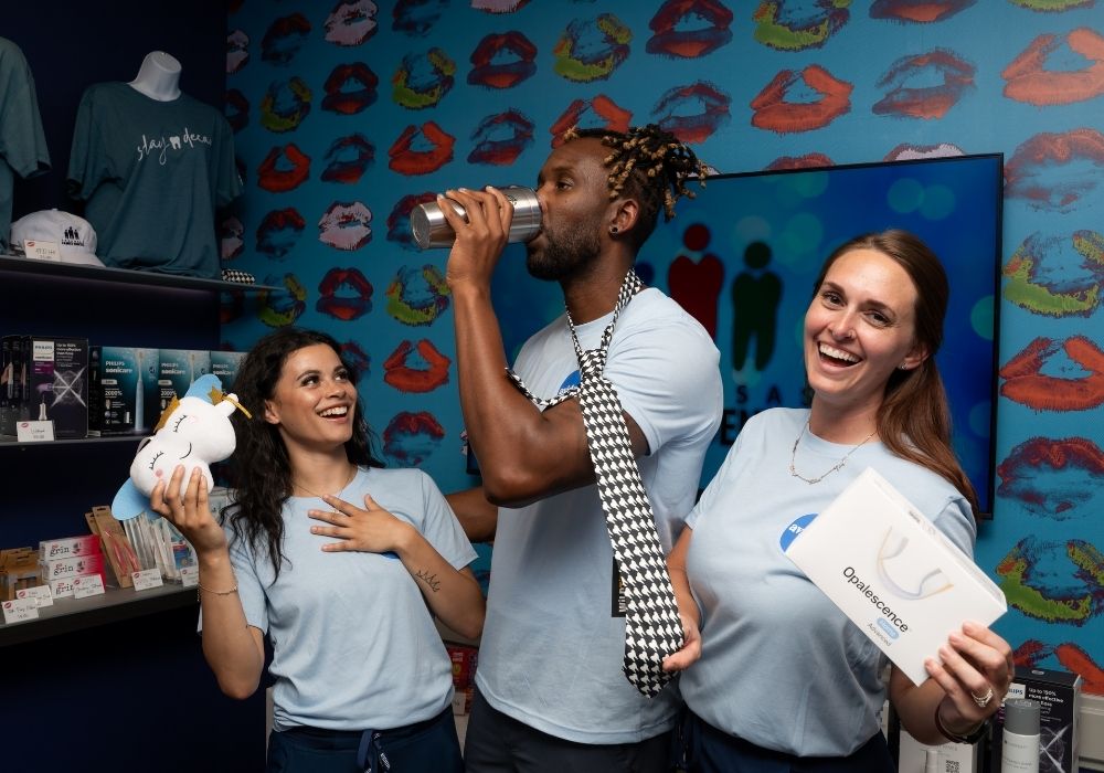 Three people in light blue shirts smile in a colorful store with lip prints on the wall. One drinks from a tumbler, another holds a plush toy, and the third holds a box labeled Opalescence..