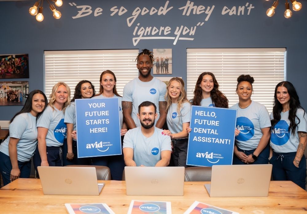 Avid Dental Home (16) A group of smiling people in matching shirts pose indoors with laptops. Two hold signs: YOUR FUTURE STARTS HERE! and HELLO, FUTURE DENTAL ASSISTANT! Inspirational quote on the wall behind them.