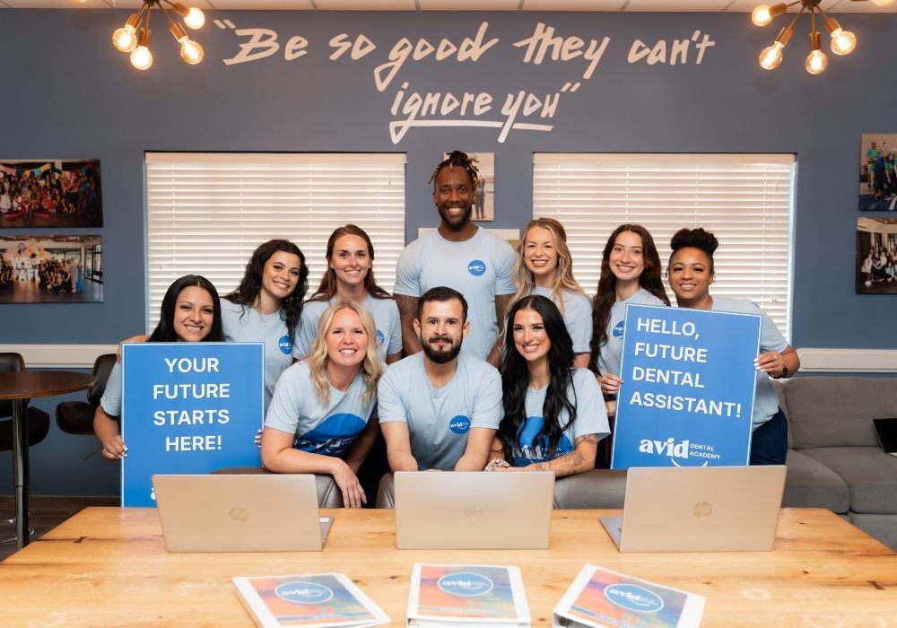 A group of smiling people in blue shirts pose indoors with laptops and signs that read YOUR FUTURE STARTS HERE! and HELLO, FUTURE DENTAL ASSISTANT! under a quote on the wall.