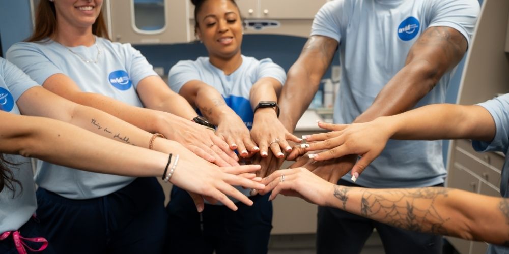 A diverse group of people in matching blue shirts join hands in the center, forming a team huddle in a brightly lit room.