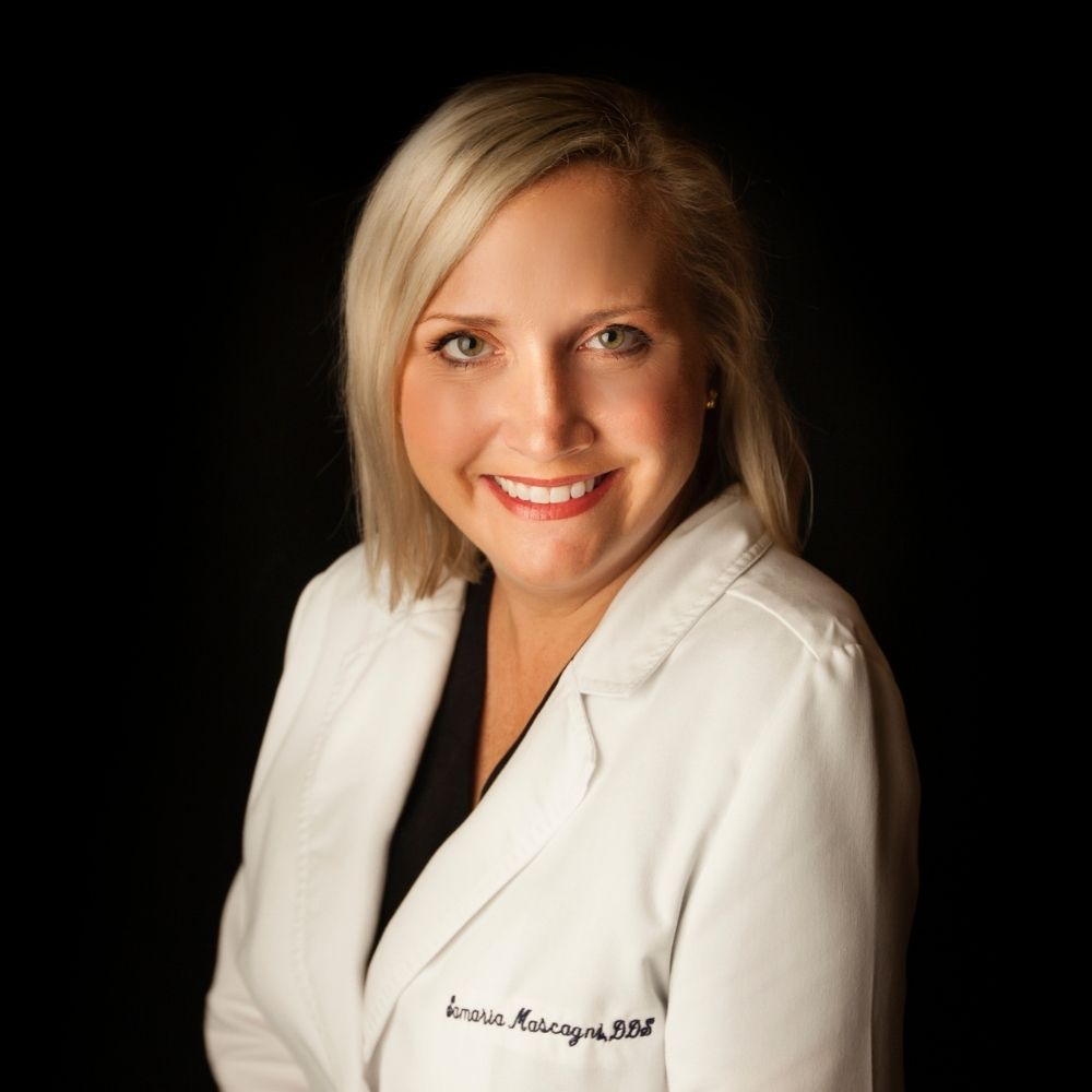 A woman with blonde hair wearing a white coat smiles against a black background. Her coat has writing on the left side.
