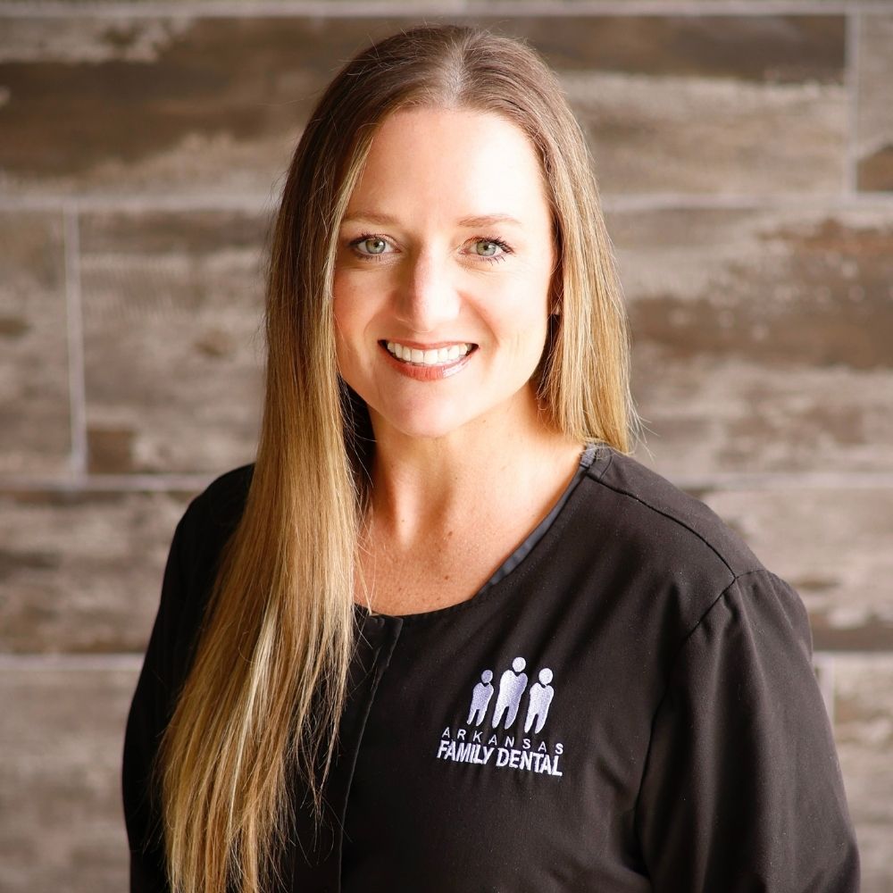 Woman with long straight hair smiling, wearing a black top that says Arkansas Family Dental, standing in front of a wooden wall background.
