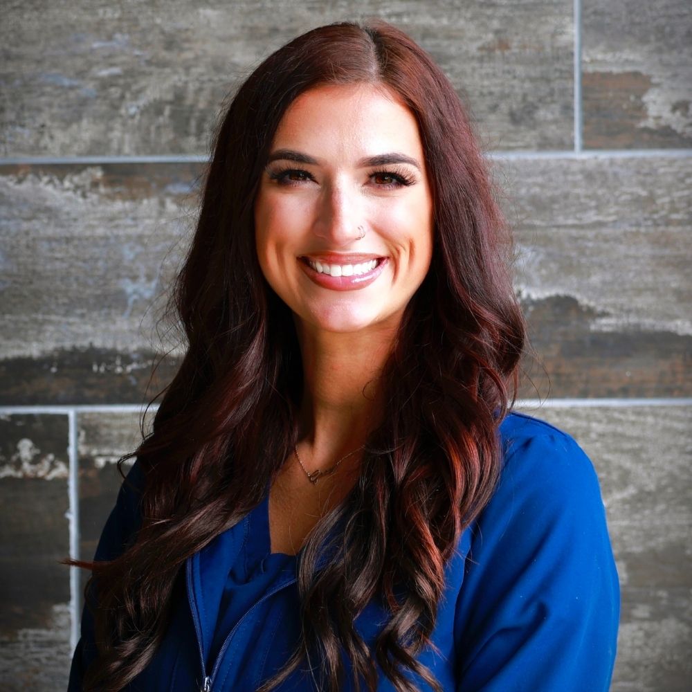 A woman with long, wavy brown hair and a blue top smiles at the camera, standing in front of a textured gray tiled wall.