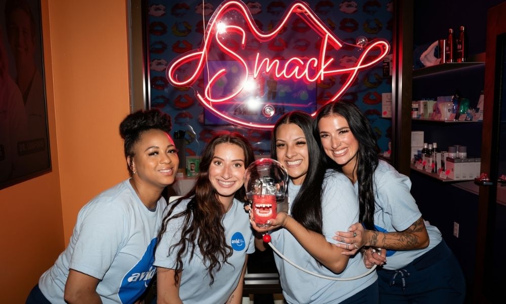 Four women in light blue uniforms smile and pose together, holding a dental model. Behind them, a neon sign shaped like lips spells Smack in cursive. The background includes colorful decor and shelves with products.
